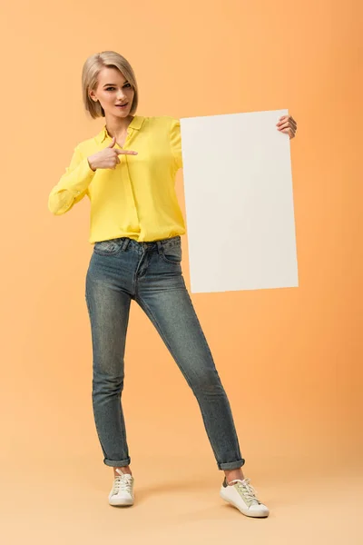 Confident blonde girl in jeans pointing with finger at blank placard on orange background — Stock Photo