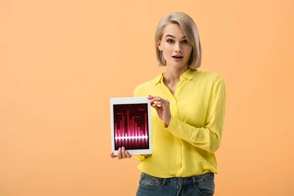 Mujer joven sorprendida sosteniendo tableta digital con aplicación de comercio en la pantalla aislada en naranja - foto de stock