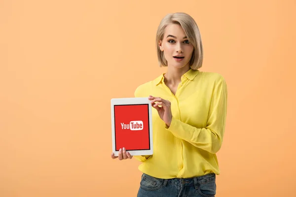 Überraschte junge Frau im gelben Hemd hält digitales Tablet mit Youtube-App auf dem Bildschirm isoliert auf orange — Stockfoto