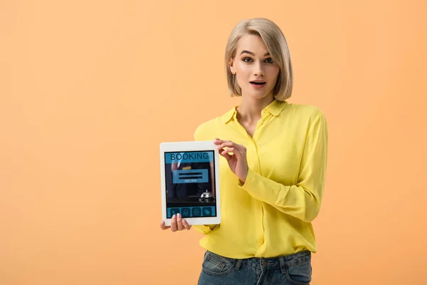 Überraschtes Mädchen im gelben Hemd hält digitales Tablet mit Buchungs-App auf dem Bildschirm isoliert auf orange — Stockfoto