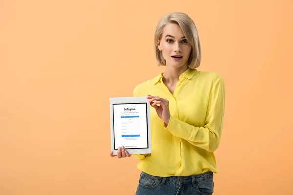 Fille surprise en chemise jaune tenant tablette numérique avec application instagram à l'écran isolé sur orange — Photo de stock