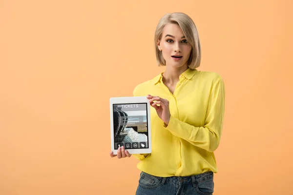 Surprise femme blonde tenant tablette numérique avec l'application de billets à l'écran isolé sur orange — Photo de stock