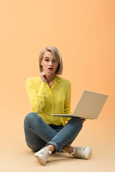 Joven rubia sorprendida en jeans sosteniendo laptop y sentada con las piernas cruzadas sobre fondo naranja - foto de stock