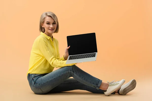 Mujer rubia sonriente en jeans mostrando portátil con pantalla en blanco mientras está sentado sobre fondo naranja - foto de stock