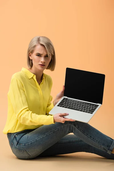 Mujer rubia trastornada en camisa amarilla mostrando portátil con pantalla en blanco sobre fondo naranja - foto de stock