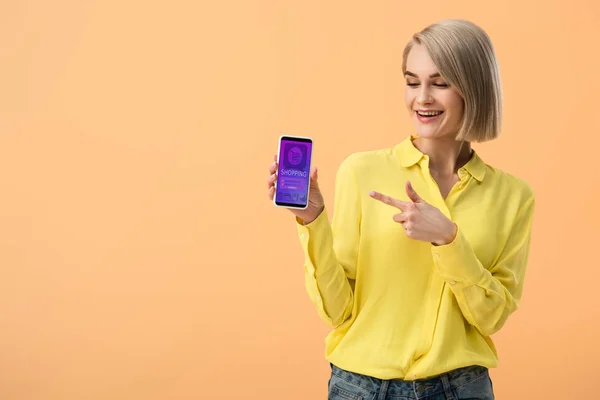 Mujer rubia sonriente apuntando con el dedo al teléfono inteligente con aplicación de compras en línea en la pantalla aislada en naranja - foto de stock