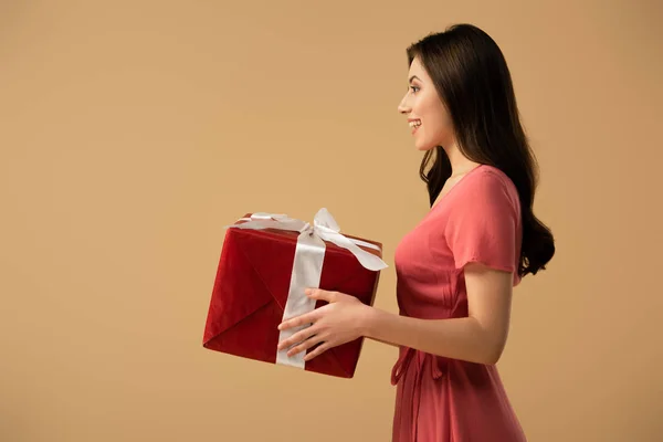 Вид сбоку счастливой брюнетки в платье с подарочной коробкой, изолированной на бежевом — стоковое фото