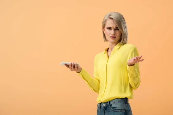 Femme blonde insatisfaite en chemise jaune tenant smartphone isolé sur orange — Photo de stock
