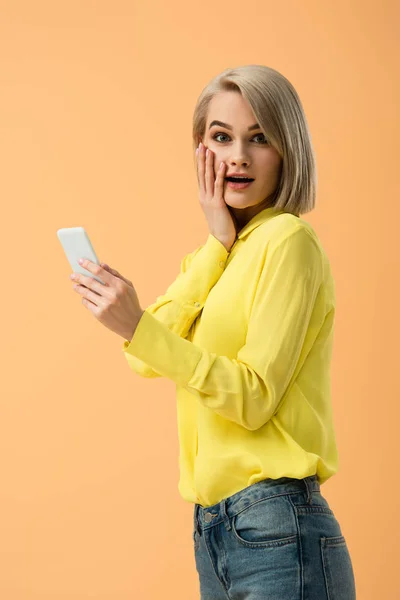 Elegante chica rubia sorprendida sosteniendo teléfono inteligente aislado en naranja - foto de stock