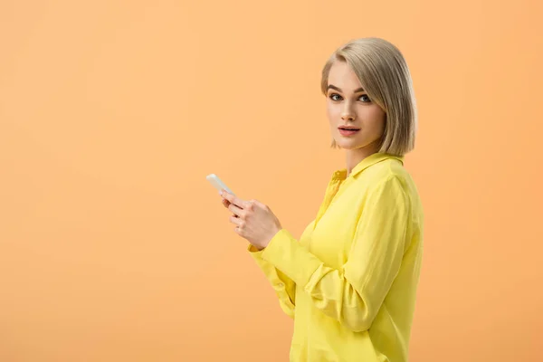 Mujer rubia bonita en camisa amarilla sosteniendo teléfono inteligente y mirando a la cámara aislada en naranja - foto de stock