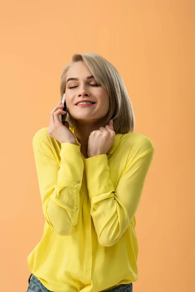 Chica rubia inspirada en camisa amarilla hablando en smartphone con los ojos cerrados aislados en naranja - foto de stock