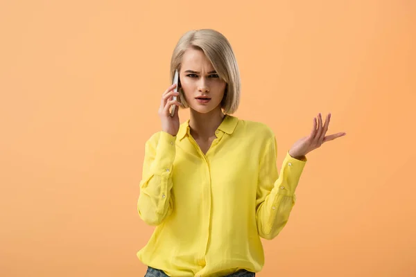 Dissatisfied blonde girl in yellow shirt talking on smartphone isolated on orange — Stock Photo