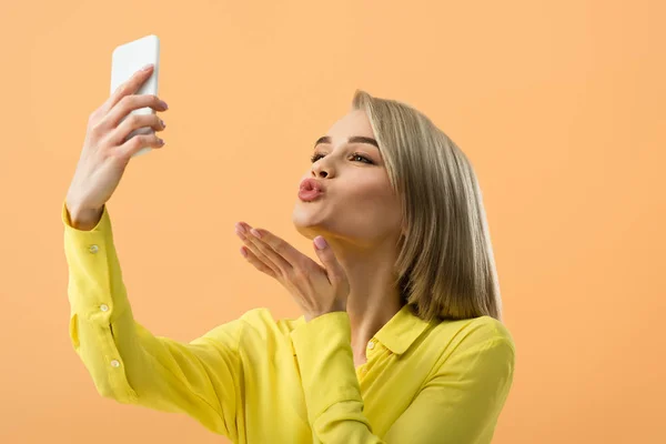 Preciosa chica rubia enviando beso de aire mientras toma selfie aislado en naranja - foto de stock