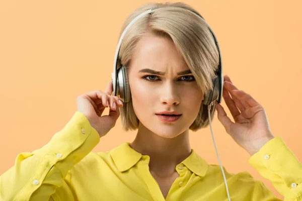 Dissatisfied blonde young woman touching headphones and looking at camera isolated on orange — Stock Photo
