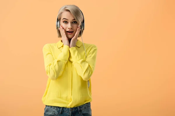 Shocked young woman in headphones touching face and looking at camera isolated on orange — Stock Photo