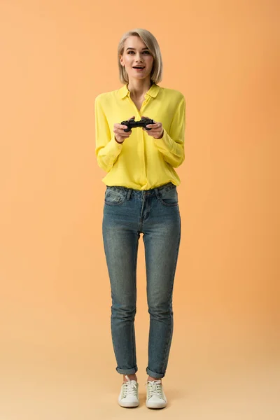 Full length view of happy blonde girl in jeans holding joystick on orange background — Stock Photo