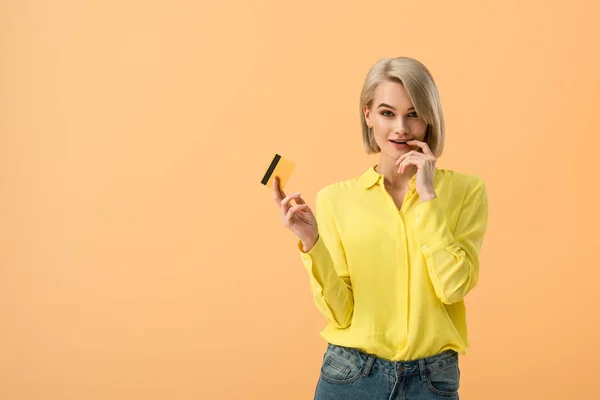 Mulher loira sensual em camisa amarela segurando cartão de crédito isolado em laranja — Fotografia de Stock