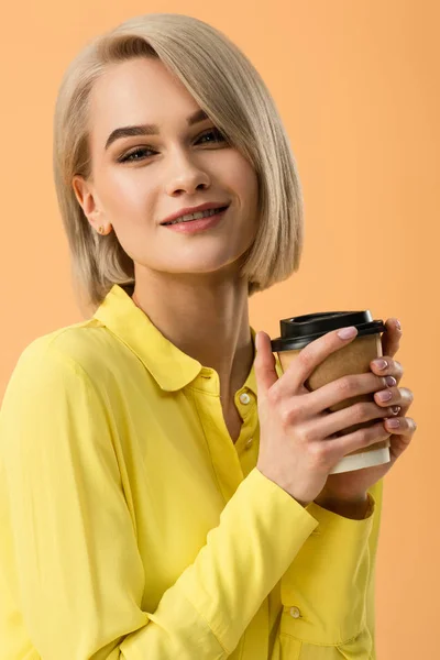 Sonriente hermosa chica rubia sosteniendo taza de papel de café aislado en naranja - foto de stock