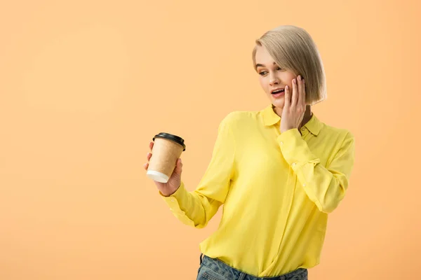 Interessato giovane donna in camicia gialla guardando tazza di carta con caffè isolato su arancione — Foto stock