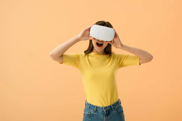 Emocionado chica morena tocando vr auriculares con la boca abierta aislado en naranja - foto de stock