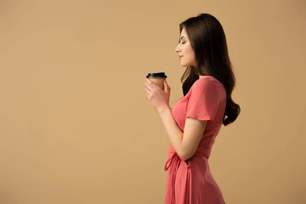 Vista laterale di elegante donna bruna che tiene la tazza di caffè isolata sul beige — Foto stock