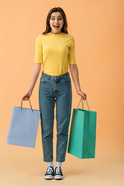 Vista completa de la chica morena sonriente sorprendida sosteniendo bolsas de compras sobre fondo naranja - foto de stock