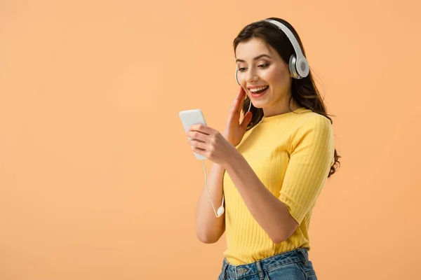 Fröhliches brünettes Mädchen in Kopfhörern mit Smartphone mit Lächeln isoliert auf orange — Stockfoto