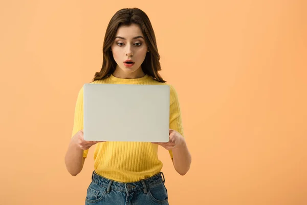 Schockierte brünette junge Frau hält Laptop isoliert auf orange — Stockfoto