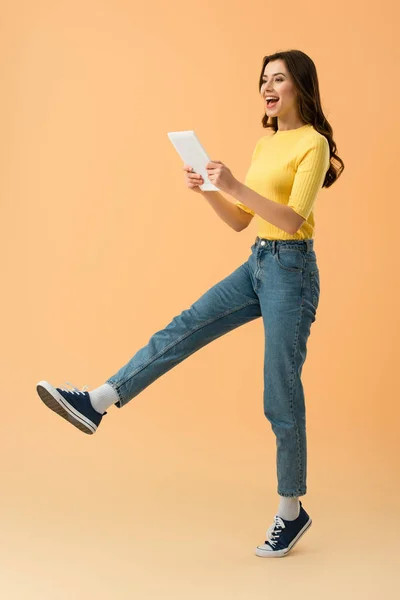 Brünettes Mädchen in Jeans hält digitales Tablet in der Hand und springt auf orangefarbenem Hintergrund — Stockfoto