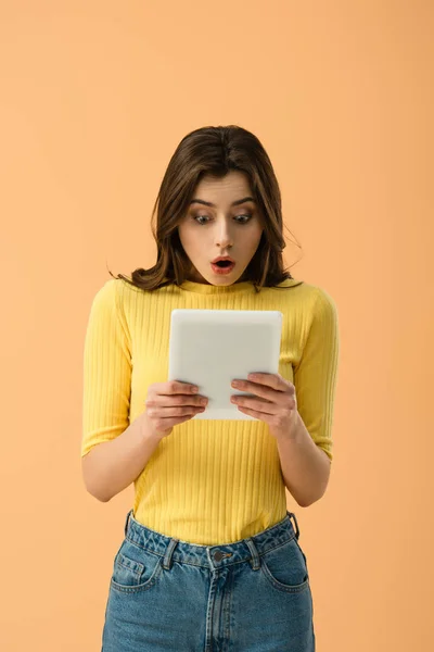 Schockierte brünette junge Frau schaut auf digitales Tablet isoliert auf orange — Stockfoto