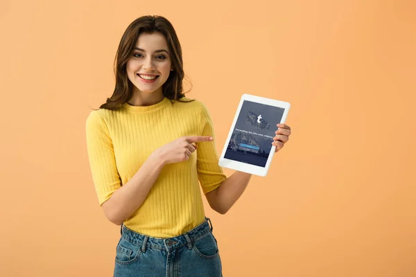 Affascinante ragazza sorridente che punta con il dito al tablet digitale con l'app tumblr sullo schermo isolato su arancione — Foto stock