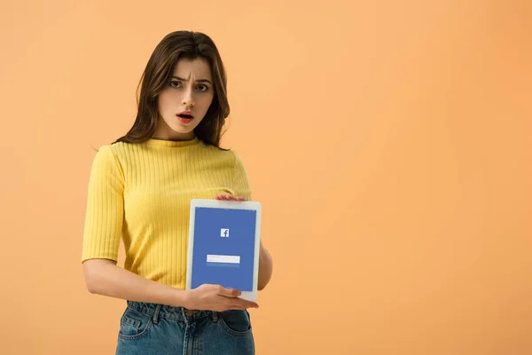 Surprised brunette young woman holding digital tablet with facebook app on screen isolated on orange — Stock Photo