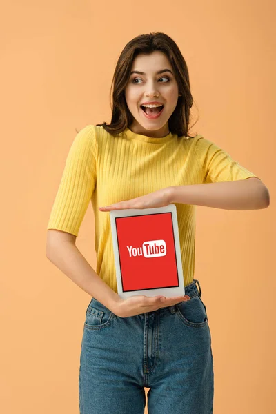 Excited brunette girl holding digital tablet with youtube app on screen isolated on orange — Stock Photo