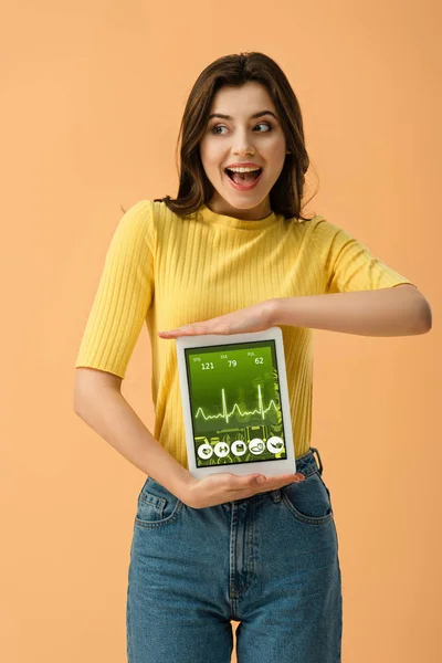 Joyful brunette woman holding digital tablet with health app on screen isolated on orange — Stock Photo