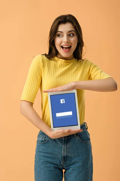 Emocionado chica morena sosteniendo tableta digital con aplicación de facebook en la pantalla aislado en naranja - foto de stock