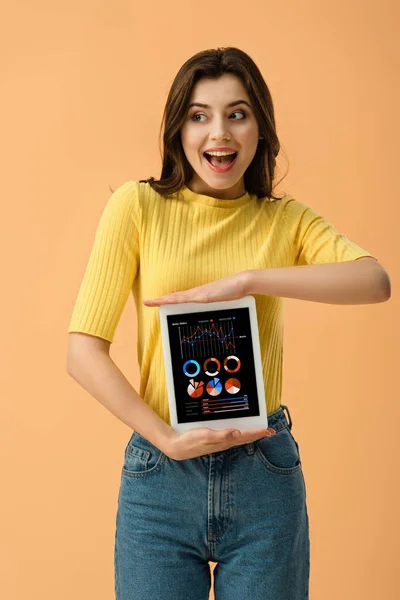 Happy brunette girl holding digital tablet with business app on screen isolated on orange — Stock Photo