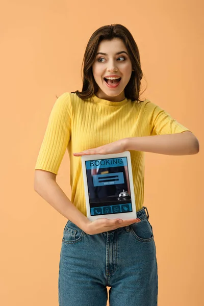 Захоплена брюнетка дівчина тримає цифровий планшет з додатком бронювання на екрані ізольовано на помаранчевому — стокове фото