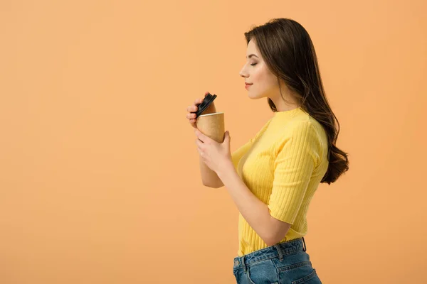 Vista lateral da mulher morena relaxada segurando xícara de papel de café isolado na laranja — Fotografia de Stock