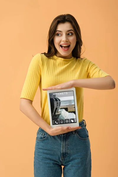 Excited brunette woman holding digital tablet with tickets app on screen isolated on orange — Stock Photo