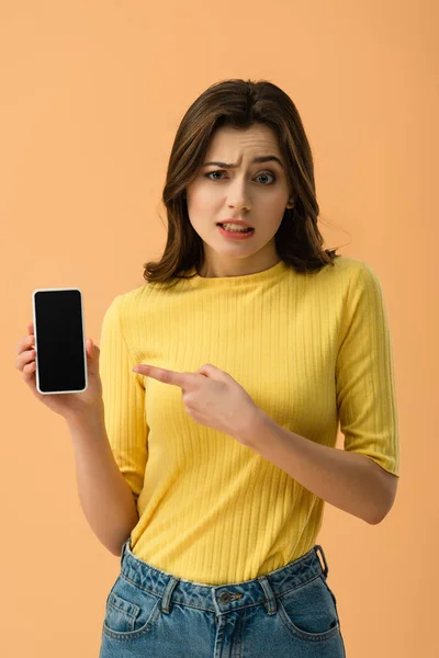 Chica morena confundida apuntando con el dedo al teléfono inteligente con pantalla en blanco aislado en naranja - foto de stock