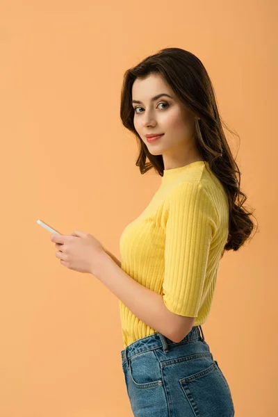 Beautiful brunette girl holding smartphone and looking at camera isolated on orange — Stock Photo