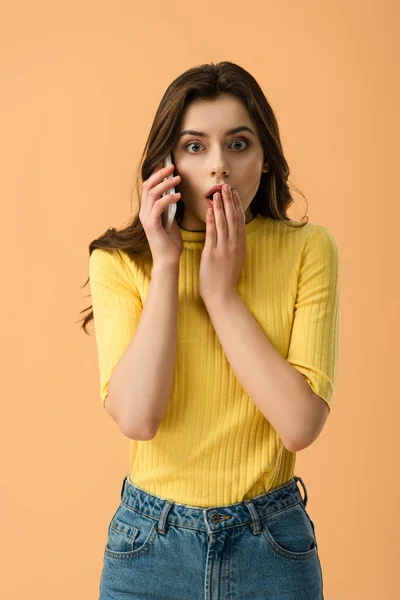 Шокированная молодая женщина разговаривает на смартфоне и закрывая рот изолирован на оранжевый — стоковое фото