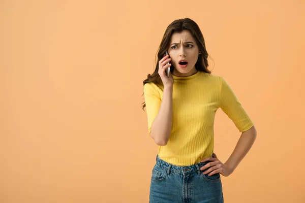Шокированная молодая женщина говорит на смартфоне, стоя с рукой на бедре изолированы на оранжевый — стоковое фото