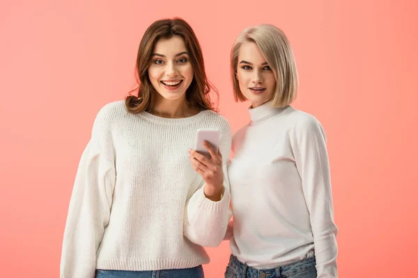 Fröhliches brünettes Mädchen hält Smartphone in der Hand und steht neben blonder Freundin isoliert auf rosa — Stockfoto