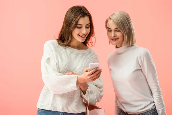 Cheerful blonde and brunette friends looking at smartphone isolated on pink — Stock Photo