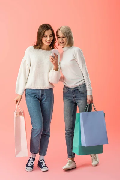 Alegre rubia y morena amigos mirando el teléfono inteligente mientras sostiene bolsas de compras sobre fondo rosa - foto de stock