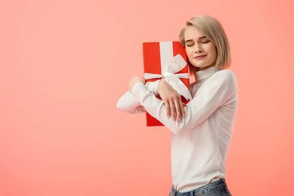 Fröhliches blondes Mädchen umarmt rote Geschenkbox isoliert auf rosa — Stockfoto