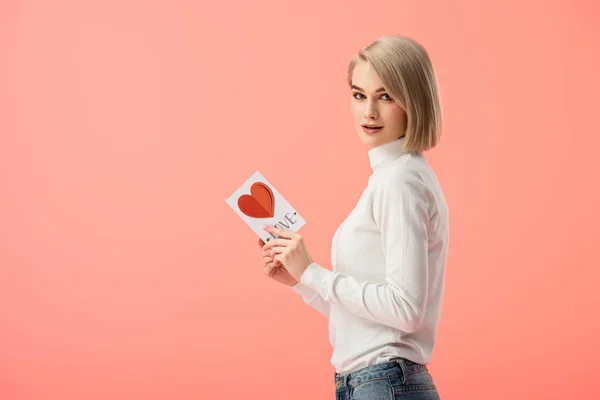 Atractiva mujer rubia sosteniendo tarjeta de felicitación aislada en rosa - foto de stock