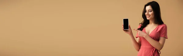 Colpo panoramico di ragazza bruna allegra che punta con il dito allo smartphone con schermo bianco isolato su marrone — Foto stock