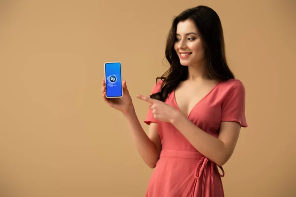 Joyeuse fille brune pointant du doigt le smartphone avec l'application shazam à l'écran isolé sur brun — Photo de stock
